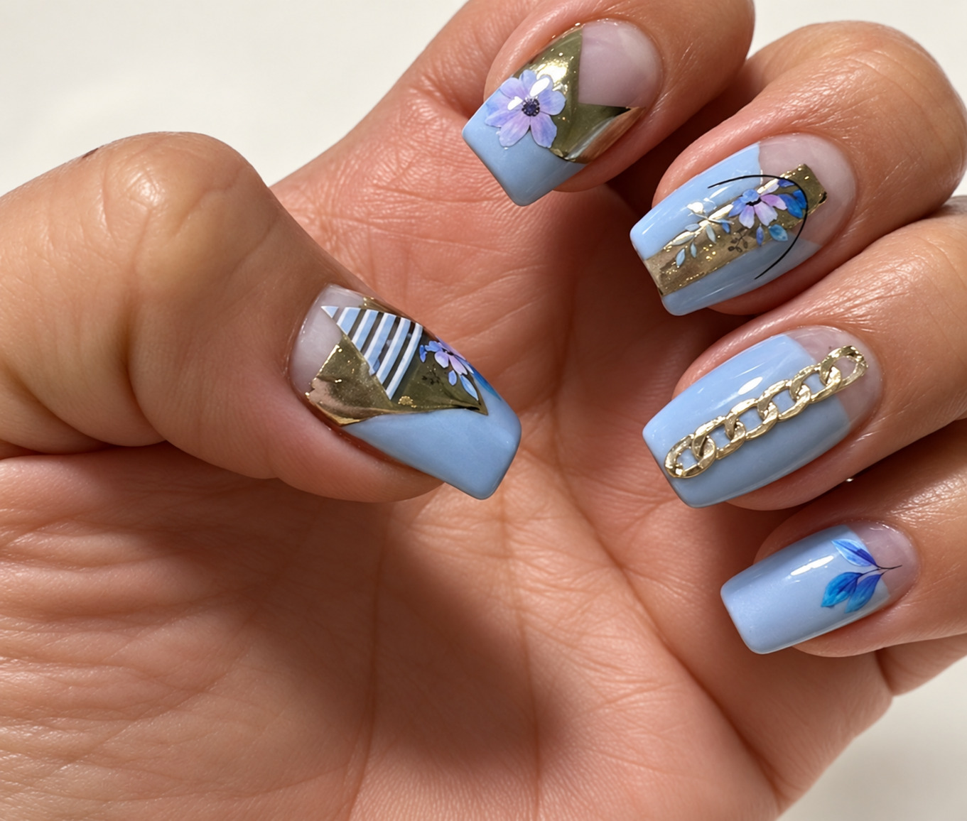 Curled hand showing lavender-blue nails with flowers, gold stripes and chain details